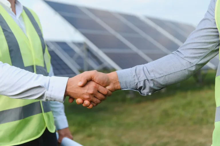 Dois técnicos em energia solar realizando um aperto de mão ao lado de placas fotovoltaicas.