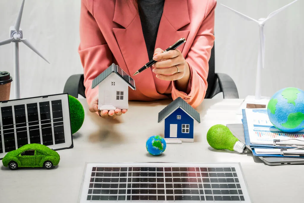 Mulher segurando uma casa em miniatura e uma caneta nas mãos, com vários paineis solares em miniatura, uma casa e um carro, também miniaturas, em cima da mesa.