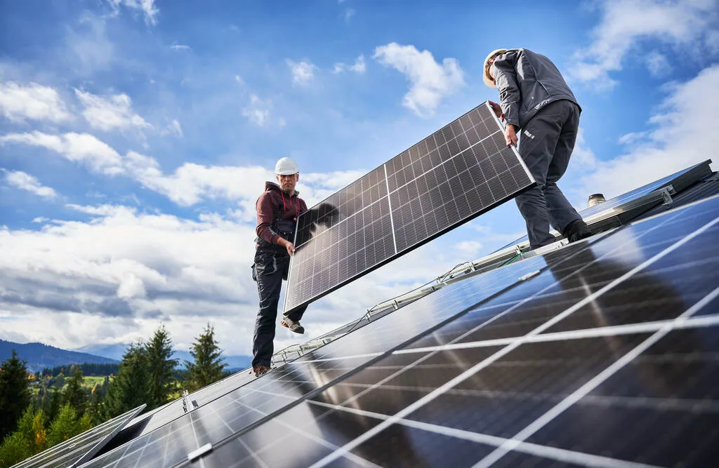 Dois trabalhadores carregando placa fotovoltaica em cima de telhado.