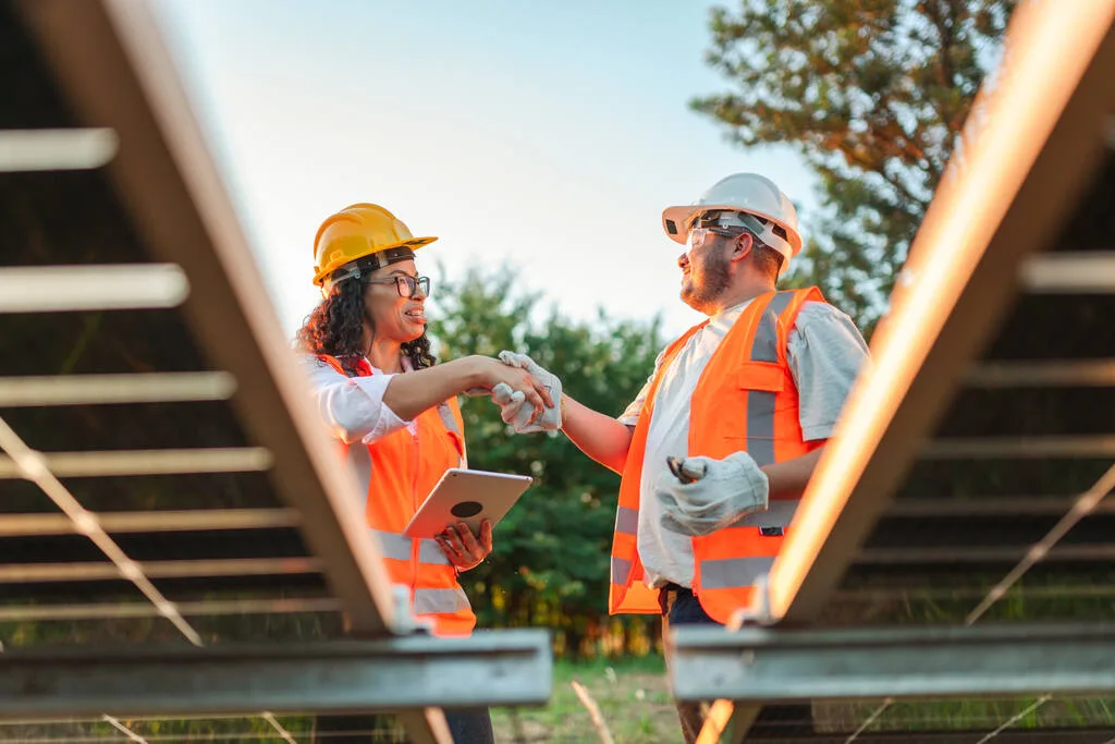 Dois trabalhadores vestidos com roupas de segurança, apertando as mãos em frente a placas solares.
