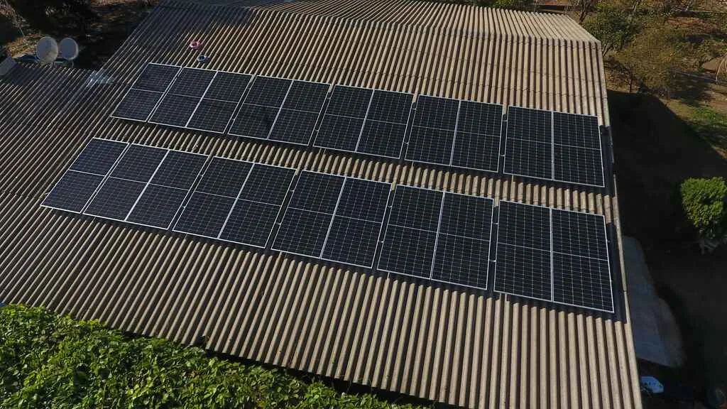 Vista aérea de um telhado grande de telhas onduladas com várias fileiras de painéis solares alinhados de forma simétrica. Os equipamentos ocupam boa parte da superfície e estão instalados em duas seções paralelas, aproveitando a incidência de luz solar para gerar energia por meio de um módulo fotovoltaico em operação residencial.