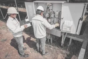 Dois homens em pé, vestindo roupas casuais e equipamentos de proteção, em frente a uma string box aberta. Um deles está com ambas as mãos na string box, enquanto o outro faz anotações em uma planilha de papel